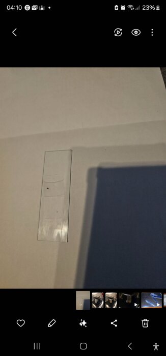 Mikroskopglas med en liten svart fläck synlig på ett ljusgrått bakgrundspapper, fotograferat ovanifrån.