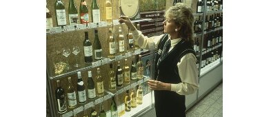 Person plockar en flaska från en hylla i en butik med vin och alkohol, omgiven av vinflaskor i olika prisklasser och sorter.