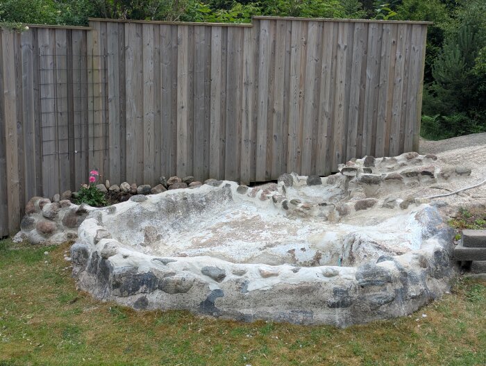 Återställd damm uppmurad med natursten längs bergssluttning framför en träplank. Gräs och en rosa blomma syns bredvid.