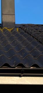 Svartmålade eternit-takpannor med flagnande färg och mossa. Synlig skorsten och klarblå himmel i bakgrunden.