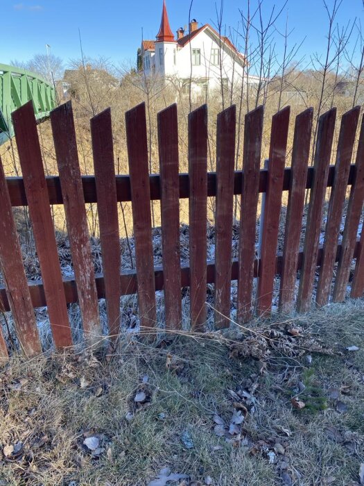 Rött staket med snedkapade toppar, framför ett vitt hus med rött tak i bakgrunden. Gräs och några torra kvistar i förgrunden.