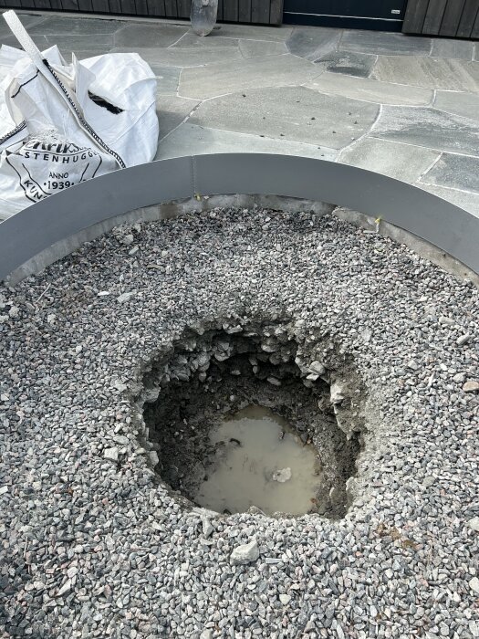 Hål i grusplan med bergkross, omgivet av en cirkel av makadam. Vatten syns i botten av hålet, redo för plantering av ett träd.