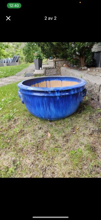 Stor blå keramikkruka med sprickor, placerad på en gräsmatta nära en terrass i trädgården.