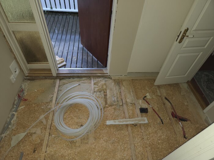 Golv med träfiberskivor och synligt rör för golvvärmeinstallation, dörrkarm och öppna dörrar, verktyg på golvet, utsikt mot veranda.