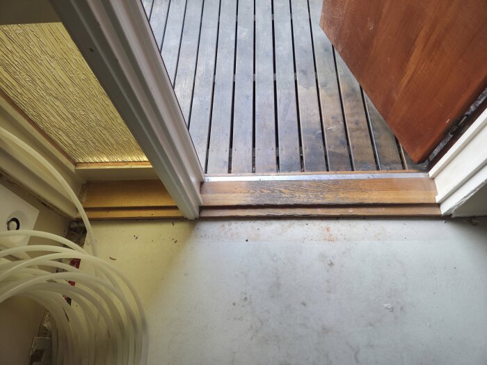 Öppen ytterdörr med synlig dörrkarm och tröskel; lite av golvet och en del av ett rullat rör syns också inomhus.