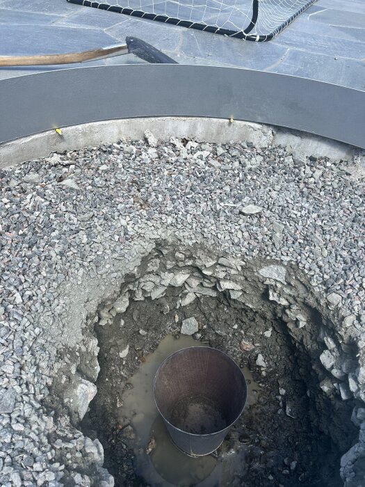 Stort hål i marken fyllt med småsten och vatten, med en kruka i mitten, omgiven av en cirkulär silverkant på stenlagd yta.