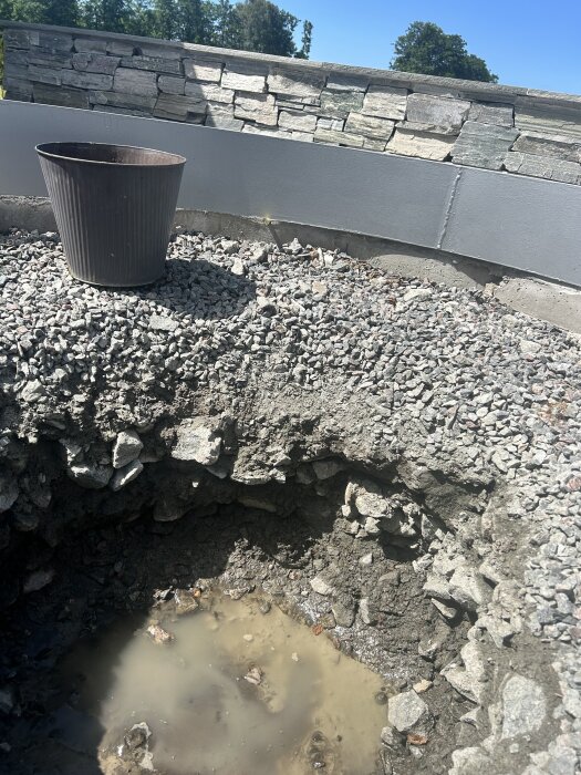 Hål i grusig mark med vatten i botten, en svart kruka bredvid. Stenmur i bakgrunden och grönska synlig ovanför muren.