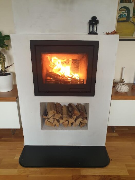 Contura i6 kamin installerad i murstock med vedutrymme under, omgiven av möbler och dekorationer.