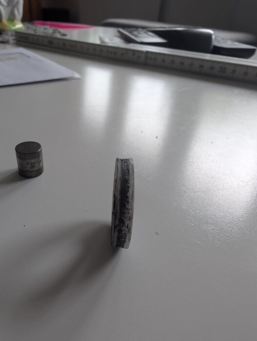 Trasigt hjul från skjutdörr, sedd från sidan på ett vitt bord, med måttband och andra föremål i bakgrunden.