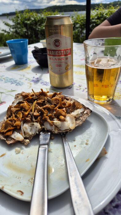 Knäckebröd med kantareller och fisk på tallrik, ölburk och glas på blommig duk utomhus, sjö och träd i bakgrunden.