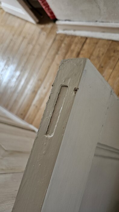 Övre vänstra hörnet av en gammal trädörr med förskjuten ram och tydlig spricka, sedd mot ett trägolv i bakgrunden.