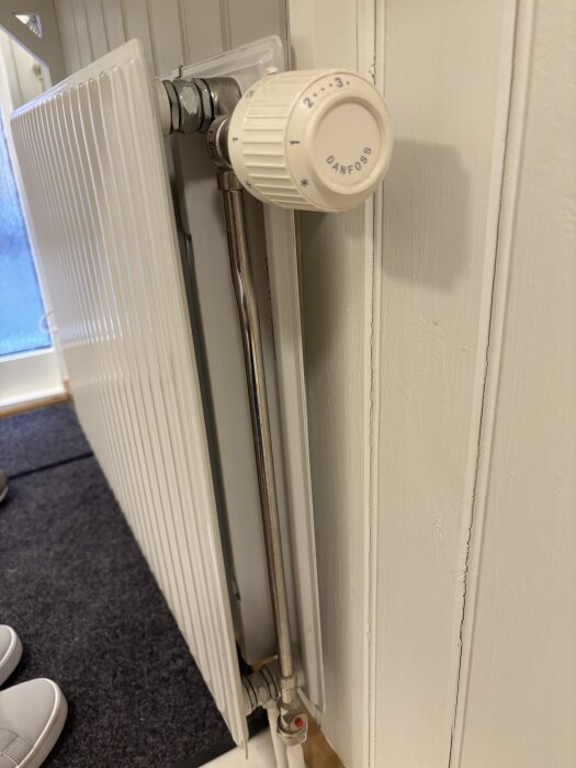 Radiator med Danfoss termostatkoppling, sett från sidan, monterad mot en vit vägg. En röd plastknopp syns på rörets nedersta del.