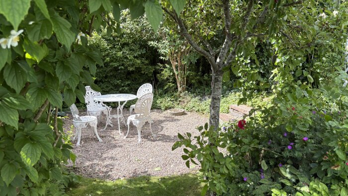 Lummig trädgård med vit trädgårdsmöbelgrupp, omgiven av tätt grönska och blommor under ett träd.