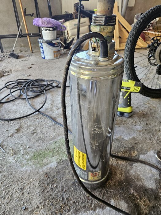 Trasig pump från en grävd brunn, uppkopplad för testning, ligger på ett golv med verktyg och cykel i bakgrunden.