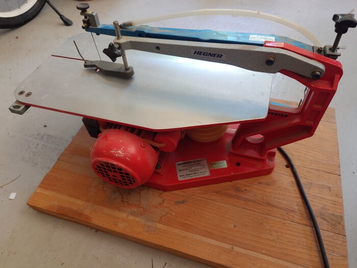 Röd Hegner kontursåg på träbord, med en metallbälg synlig. Verktyget är oanvänt men gammalt och behöver kanske en reservdel.