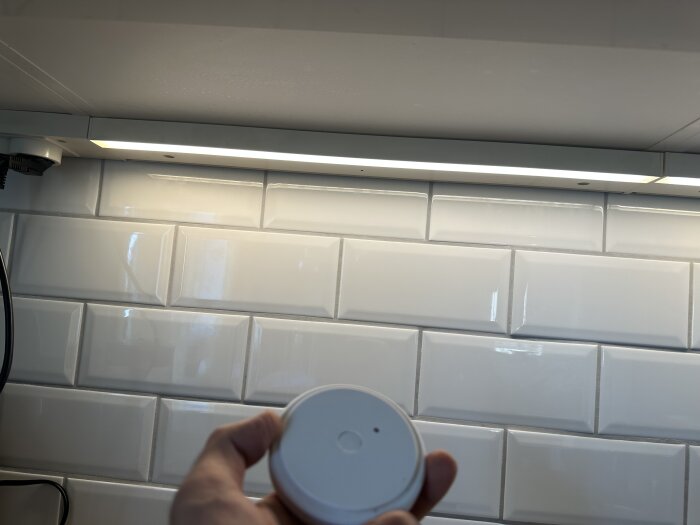 Hand håller fjärrkontroll framför en tänd LED-lampa under ett köksskåp med vit kakelvägg i bakgrunden.