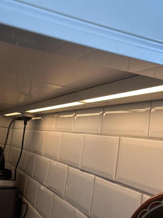 Underskåpsbelysning med en lysande LED-lampa i köket, vit kakelvägg synlig i bakgrunden.