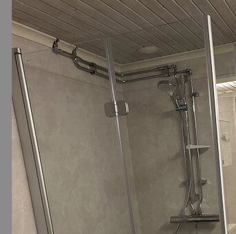 Väggmonterade vattenrör i badrum, delvis synliga längs taket, ansluter till duscharmatur. Duschen flyttad, rören dragna runt hörnet enligt kontexten.