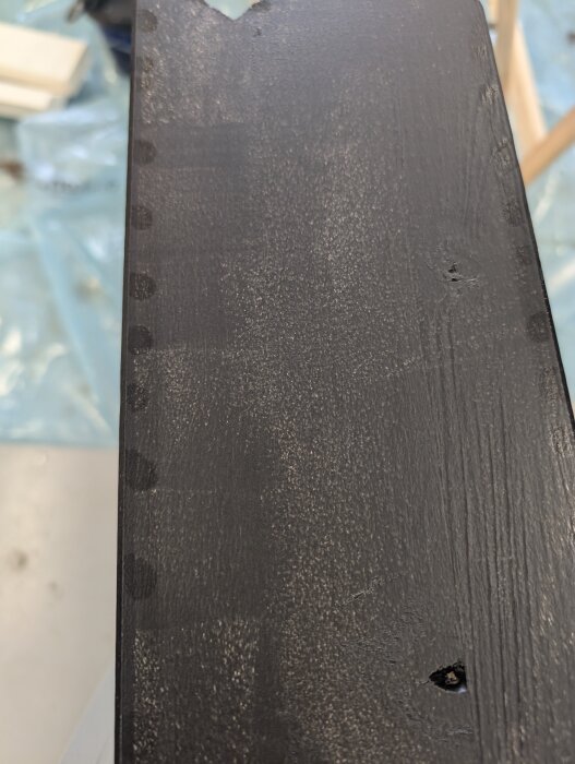 Svartmålat trästycke med klibbiga fläckar längs kanterna, grundat med oljebaserad primer och täckt med fasadfärg, torkat i varmgarage.