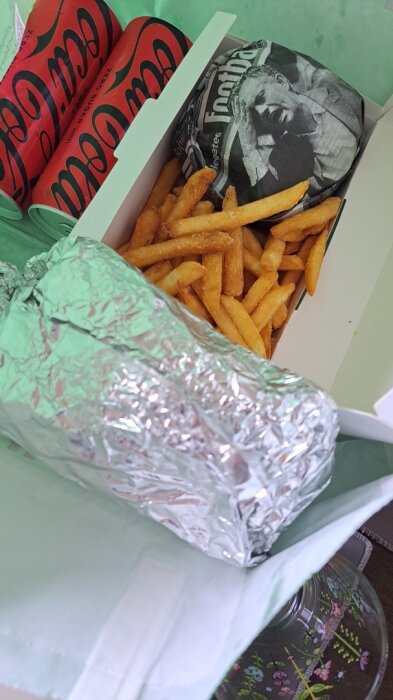 Lammkorvsrulle i folie med pommes frites och läskburkar i en kartong.