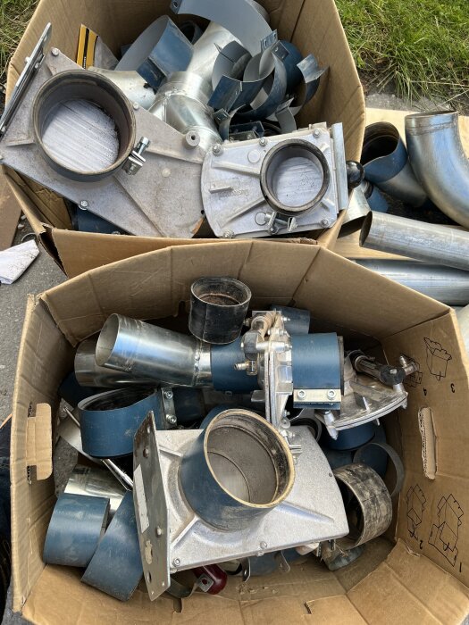 Kartonger fyllda med metallrör och kopplingar till ett spånsuganläggningsprojekt, inklusive spjäll och anslutningsdetaljer.