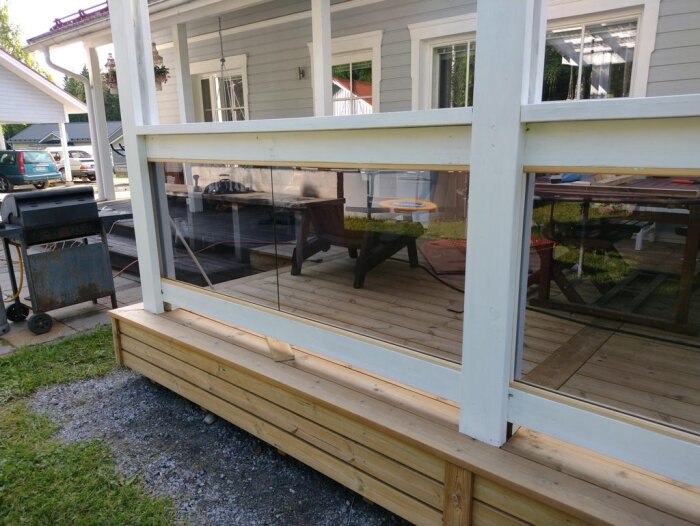 Ingång till veranda med glasskivor monterade mellan vita trästolpar, en grill och bilar syns i bakgrunden.