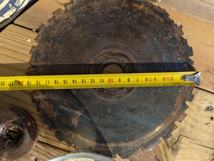 Rostig grästrimmerklinga mäts med ett måttband för att kontrollera håldiameter, uppmätt till cirka 25 mm, på ett träbord.