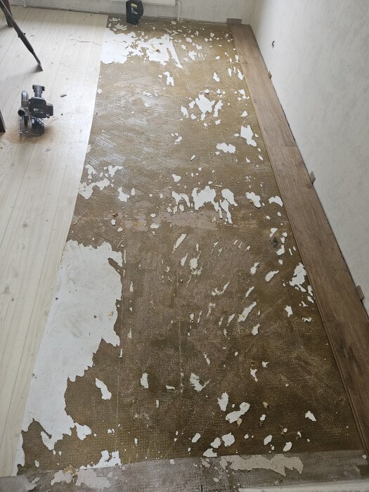 Borttagen plastmatta avslöjar limrester på golvet, med påbörjad ny golvläggning längs kanten och en verktygsmaskin synlig till vänster.