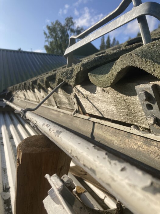 En närbild av en takfot med takpannor och en planka under, samt en takränna. Trä är synligt med tecken på fukt och möjlig röta.