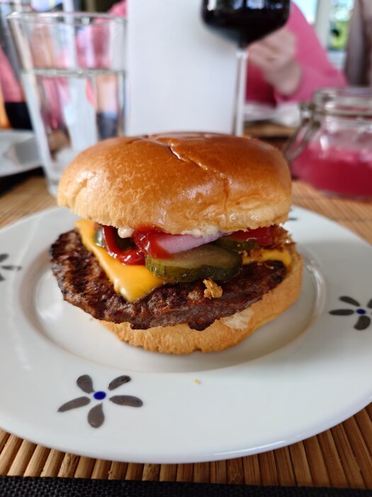 Hamburgare med ost, ketchup, rostad lök, picklad lök, saltgurka och senap på ett fat med blommotiv.