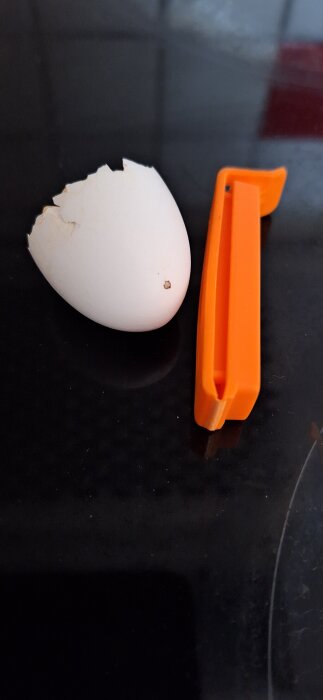 Vitt sprucket äggskal bredvid ett orange plastföremål på en svart yta.