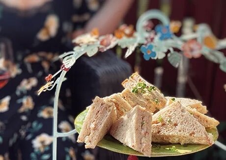 Smörgåsar med olika fyllningar på ett grönt fat, dekorerade med grönska och placerade på ett bordsställ med blommiga detaljer.