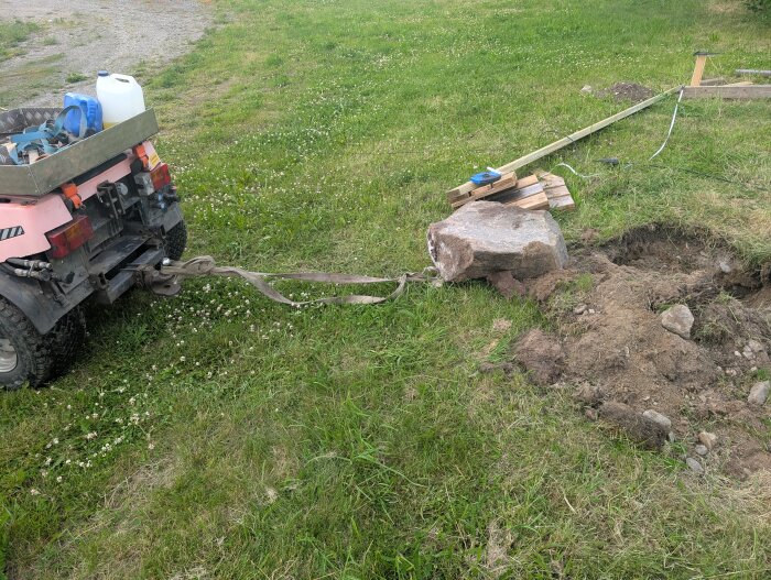 Traktor drar upp en stor sten från gräsmatta med rem, omgiven av jord och brädbitar, vid byggandet av en kaninbur.