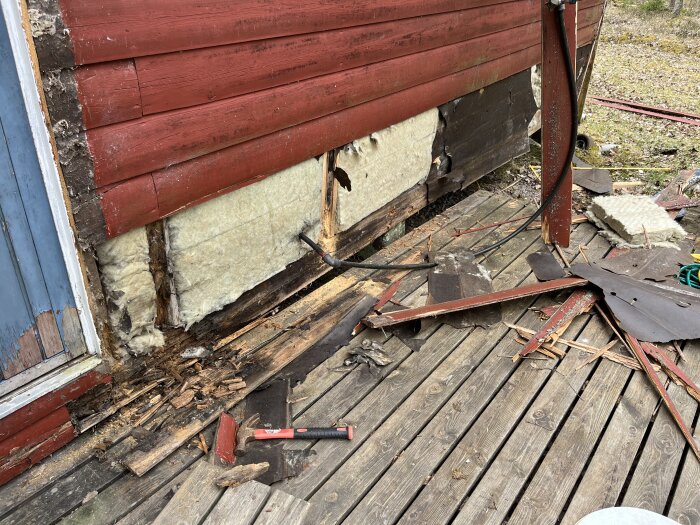 Röd stuga med synlig isolering och trasigt trä vid botten. Verktyg och träspill på träaltan. Renoveringsprojekt pågår vid husgrund.
