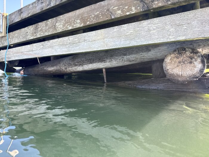 Träbrygga med slitna och ruttna stockar som ligger ojämnt över vatten, med stenkonstruktion delvis synlig under ytan.