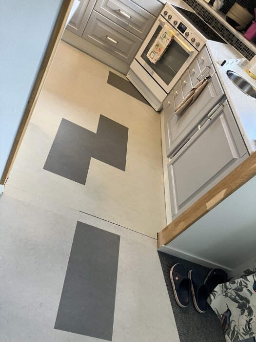 Kök med nylagt golv i ljusgrått och mörkgrått mönster; saknar tröskel och golvlist. Grå köksskåp och ugn syns, samt svarta tofflor vid väggen.