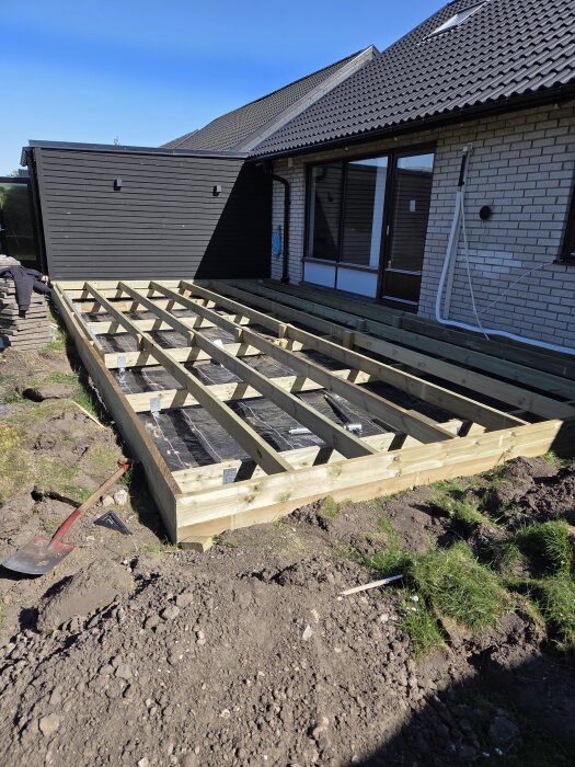 Byggandet av ett trädäck i trä utanför ett hus med nylagt ramverk, liggandes på en grusig yta med uppgrävd jord runt omkring.
