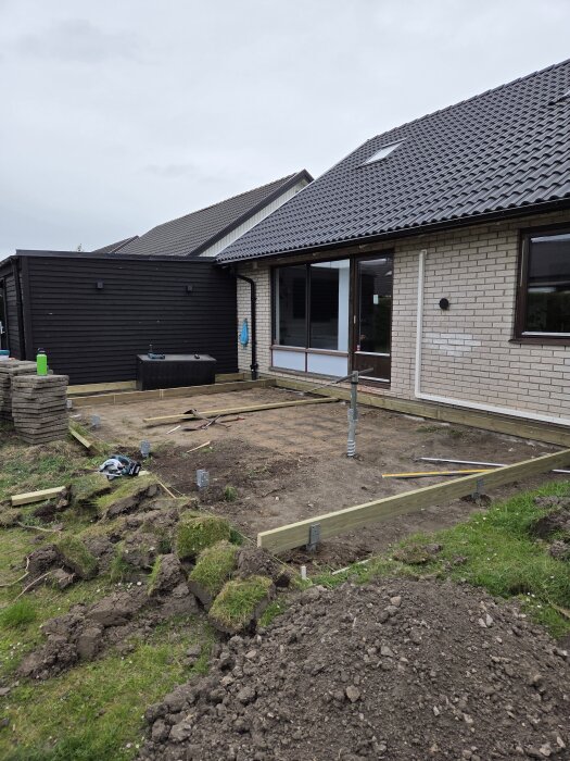 Bygge av trädäck och utekök under konstruktion; plankor, byggmaterial och jord syns bredvid ett hus med mörk och ljus exteriör.
