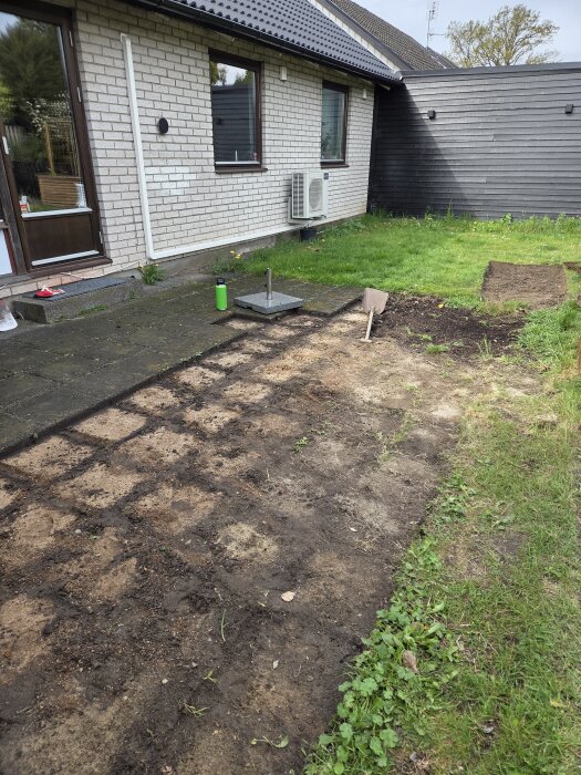 Trädgård med förberett område för trädäck och utekök vid husvägg, spade och föremål på marken. Gräsmatta och stenlagd yta syns.
