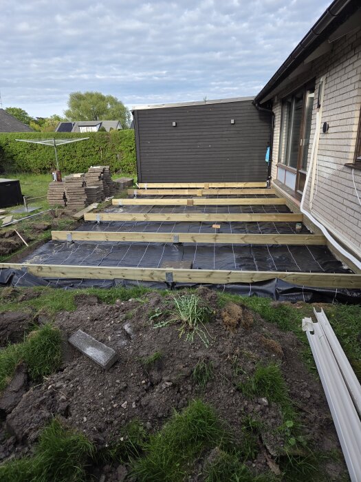 Byggnation av ett större trädäck med ramverk och markförberedelser vid ett hus, trädgård med staplade stenar och grönska i bakgrunden.