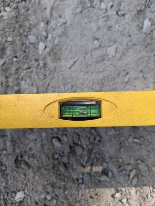 Vattenpass placerat på grusig mark visar en plan yta innan justering av ett rör med 1 cm för växthusets grund.