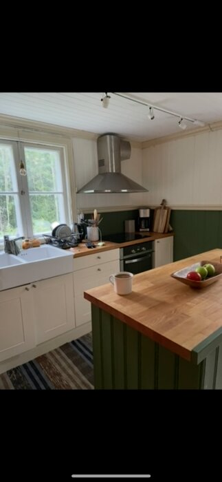 Kök med fönster, vit diskbänk, bänkskivor i trä, modern ugn och fläkt, grön panel och fruktskål på köksön.