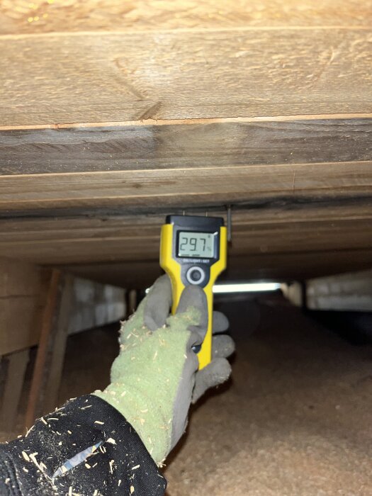 En person med handske använder en fuktmätare på träreglar i en vind, mätaren visar 29,7.