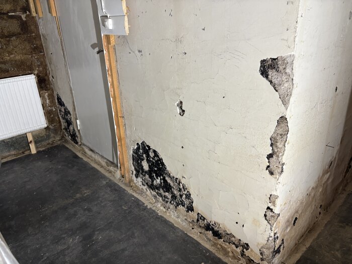 Vägg i källare med vit puts och svart massa bakom, vissa delar av putsen har lossnat och visar den svarta massan, potentiellt asbestinnehåll.