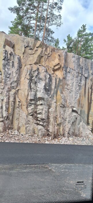 Bergvägg med tydliga sprickor och skrovlig yta, omgiven av träd på toppen och asfaltkanten nedanför.