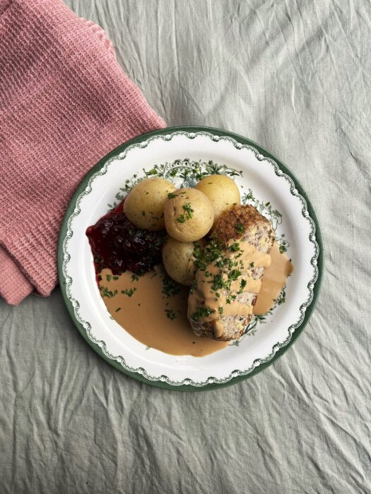 Tallrik med köttfärslimpa i sås, kokt potatis och lingonsylt, på en duk med en rosa handduk vid sidan.