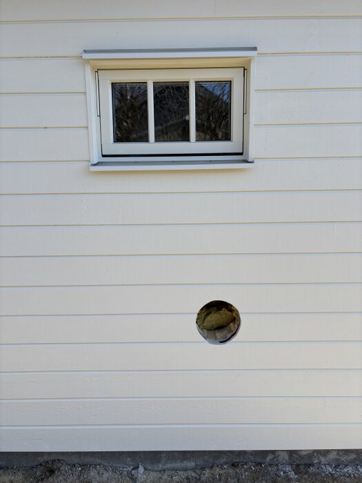 Vit vägg med ett runt hål för ventilation under ett litet fönster.