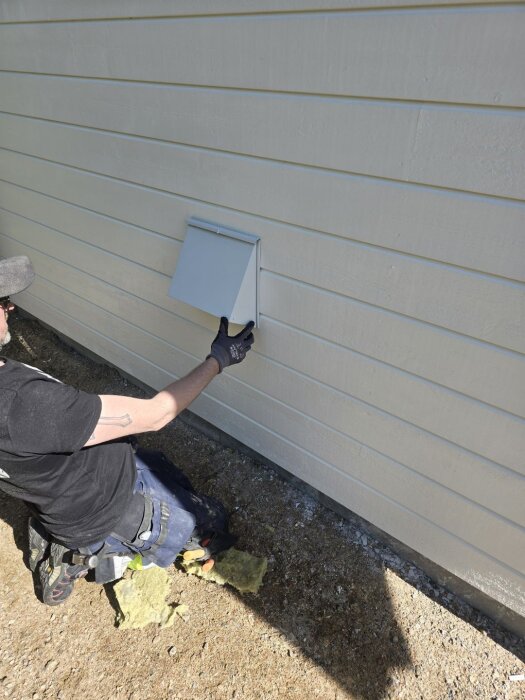 En person installerar en diskret plåtkåpa på utsidan av en byggnadsvägg, del av ventilation med brandspjäll och kallrasskydd.