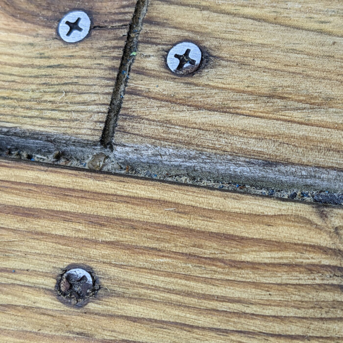 Närbild av en träyta med skruvar och sprickor, där ytan ser behandlad ut men sprickorna verkar torra. Fråga om behandlingen är med såpa eller olja.