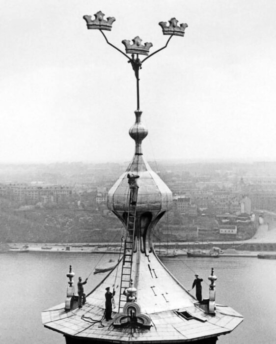 Svartvit bild av tre arbetare i arbete uppe på tornet av Stockholms stadshus, en klättrandes bland tre kronor högst upp.